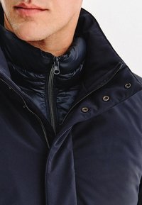 Granatowa kurtka puchowa z wysokim kołnierzem, z suwakiem z przodu i guzikami jako akcentami. Gładka tekstura z pikowanym wnętrzem.