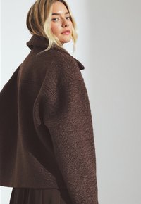 Una donna bionda guarda oltre la spalla indossando un giubbotto oversized marrone scuro strutturato su uno sfondo chiaro e neutro.