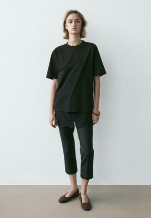 Femme debout avec une expression neutre, portant un t-shirt noir ample, un pantalon noir court, une sous-couche en dentelle, des ballerines marron et des bracelets au poignet droit.