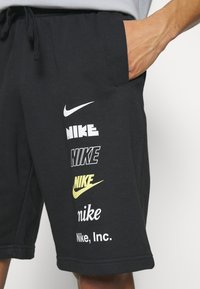 Personne portant un short noir avec plusieurs logos Nike et du texte en blanc et jaune sur la cuisse gauche, la main reposant dans la poche.