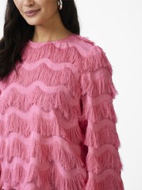 Pink top with a wavy fringe texture, featuring sheer fabric panels and a round neckline. Accented with long sleeves and soft fraying edges.