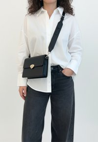 Borsa a tracolla in pelle nera con chiusura in metallo dorato, design rettangolare e tracolla regolabile, indossata sopra una camicia bianca e jeans scuri.