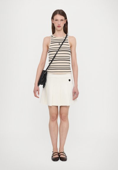 Striped tank top in cream and black, paired with a white pleated skirt featuring a logo detail; model carrying a black fringe bag and wearing brown flats.