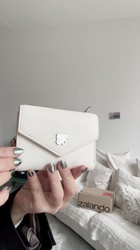 Weißes strukturiertes Lederportemonnaie mit einer dreieckigen Klappe und einem glänzenden Metalllogo. Auf der Seite ist eine sichtbare Reißverschlusstasche.