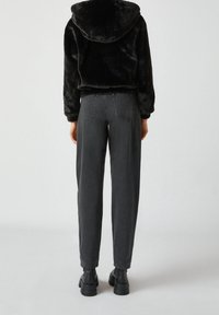 Veste en fausse fourrure noire avec capuche, associée à un jean gris taille haute. Le modèle porte des bottes noires épaisses. Vue arrière sans motifs visibles.