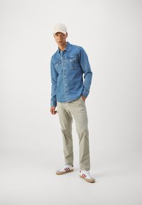 Camicia di jeans con bottone, di colore azzurro chiaro, due tasche sul petto, abbinata a pantaloni beige e scarpe bianche con strisce rosse.