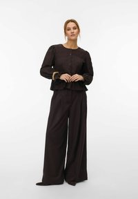 Kvinne iført en knappet, strukturert svart bluse med lange ermer, volangmansjetter og vide svarte bukser, stående mot en ensfarget hvit bakgrunn.