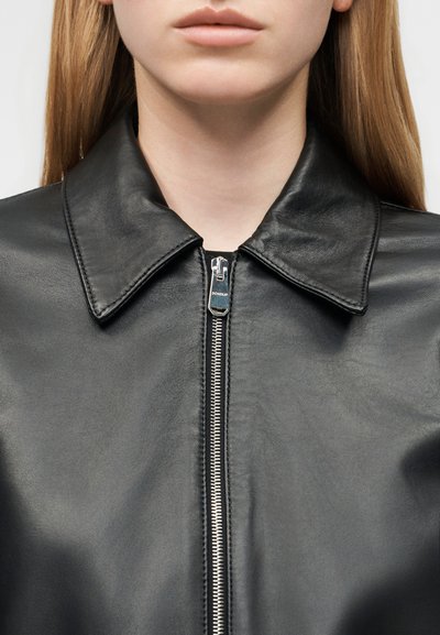 Γυναίκα με μακριά ξανθά μαλλιά φοράει μαύρο δερμάτινο μπουφάν με φερμουάρ και μυτερό γιακά, εμφανιζόμενη από τον λαιμό έως το στήθος.