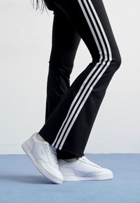 Svarta träningsbyxor med tre vita ränder på sidorna, tillsammans med vita sneakers med subtila designelement, på en blå yta.