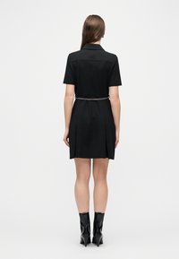 Μια γυναίκα στέκεται πλάτη, φοράει κοντό μαύρο φόρεμα με ασημένια ζώνη αλυσίδα και μαύρες μποτάκια αστραγάλου με τακούνια σε απλό λευκό φόντο.