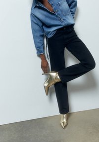 Personne portant une chemise en denim bleu et un pantalon noir, levant une jambe pour montrer des bottines à talons hauts dorées contre un mur blanc.