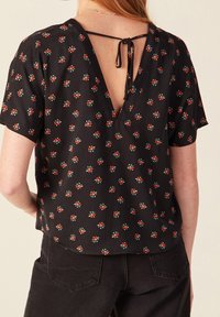 Donna che indossa una blusa nera a maniche corte con un piccolo motivo floreale rosso e una profonda scollatura a V sul retro legata con un sottile spago nero.