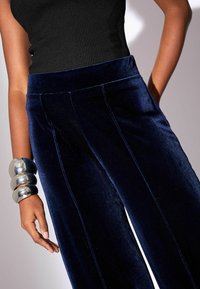 Námornícky modré sametové nohavice s širokým nohavicami a vysokým pásom, s prednými švami a hladkou textúrou. Strieborné náramky na zápästí.