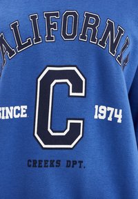 Sudadera azul con texto impreso en blanco y marino: "CALIFORNIA," "C," "1974," y "CREEKS DPT." Diseño de cuello redondo con una textura suave.