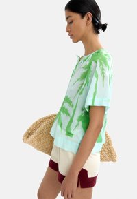 Jasnozielony top z krótkim rękawem z nadrukiem palm, połączony z kremowymi i burgundowymi szortami. Modelka trzyma plecioną torbę ze słomy.