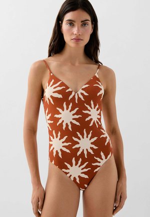 Mujer con bañador de una pieza color óxido con patrón de rayos de sol blanco y tirantes finos, de pie frente a un fondo liso y claro.