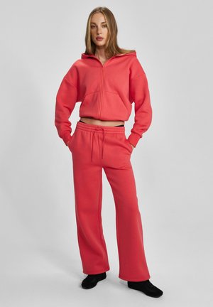 Kobieta stojąca z rękami w kieszeniach, ubrana w czerwoną bluzę rozpinaną na zamek oraz pasujące szerokie spodnie, na nogach czarne buty, na jednolitym tle.