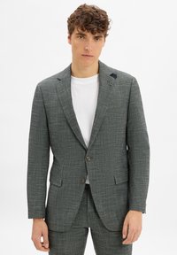 Grau karierter Blazer mit Reverskragen, einreihiger Knopfverschluss und passenden Hosen. Über einem weißen T-Shirt mit subtiler Textur getragen.