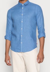 Camisa de lino azul claro con mangas largas arremangadas, cuello botón y botones visibles, que presenta un logo sutil en la parte inferior derecha.