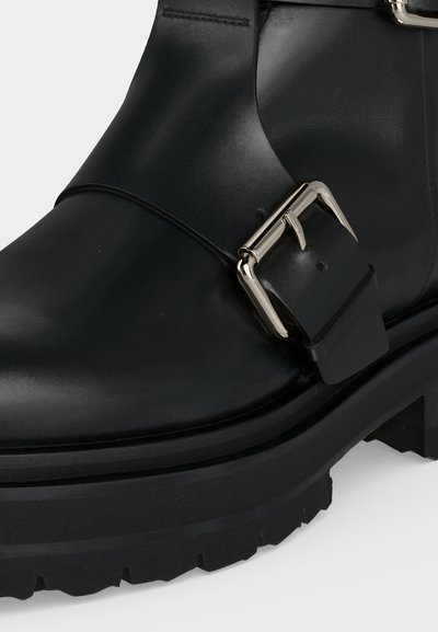 Gros plan d'une botte en cuir noire avec une semelle épaisse et une boucle rectangulaire argentée sur le côté.