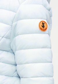 Μανίκι ανοιχτό μπλε γαλάζιας καπιτονέ μπουφάν με πορτοκαλί κυκλικό έμβλημα που απεικονίζει μια μαύρη πάπια και το κείμενο "SAVE THE DUCK".