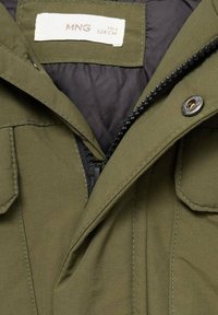 Giacca verde oliva con zip nera, bottoni a pressione, due tasche a pattina e etichetta interna che mostra la taglia 8 e 128 cm.