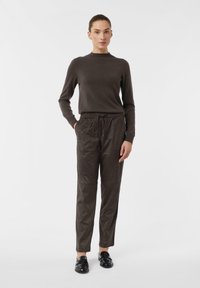 Woman standing with hands in pockets, wearing dark brown long-sleeve top, matching pants, and black loafers on a white background.