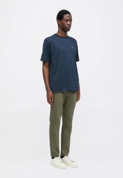 Mann, der vor einem weißen Hintergrund steht und ein navyblaues T-Shirt, olivgrüne Hosen, schwarze Socken und weiße Sneaker mit Schnürsenkeln trägt.