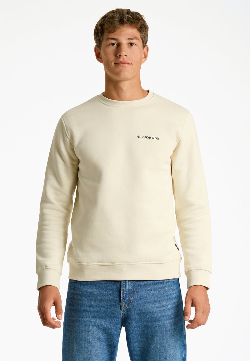Sudadera crema con cuello redondo, puños y dobladillo de canalé. Presenta un pequeño logo negro "JUNK de LUXE" en el lado izquierdo del pecho. Combinada con jeans azules.