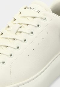 Ténis de couro branco com atacadores planos, perfurações subtis na lateral e ilhóses de cor clara. Textura suave com design minimalista.