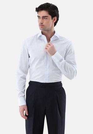 Mann richtet den Kragen eines weißen, langärmeligen Hemdes mit dünnen blauen Streifen, trägt schwarze plissierte Hosen und steht vor einem schlichten Hintergrund.