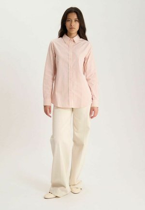 Femme debout portant une chemise rose rayée boutonnée, un pantalon crème à jambes larges et des chaussures crème sur un fond uni.