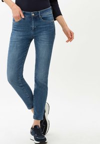 Blaue Denim-Skinny-Jeans mit hoher Taille, vorderen Taschen und Reißverschlussdetails, kombiniert mit schwarzen und weißen Sportschuhen.
