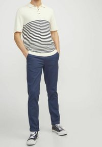 Man wearing a cream polo shirt with navy stripes, navy slim-fit pants, and black and white sneakers, standing with hands in pockets.