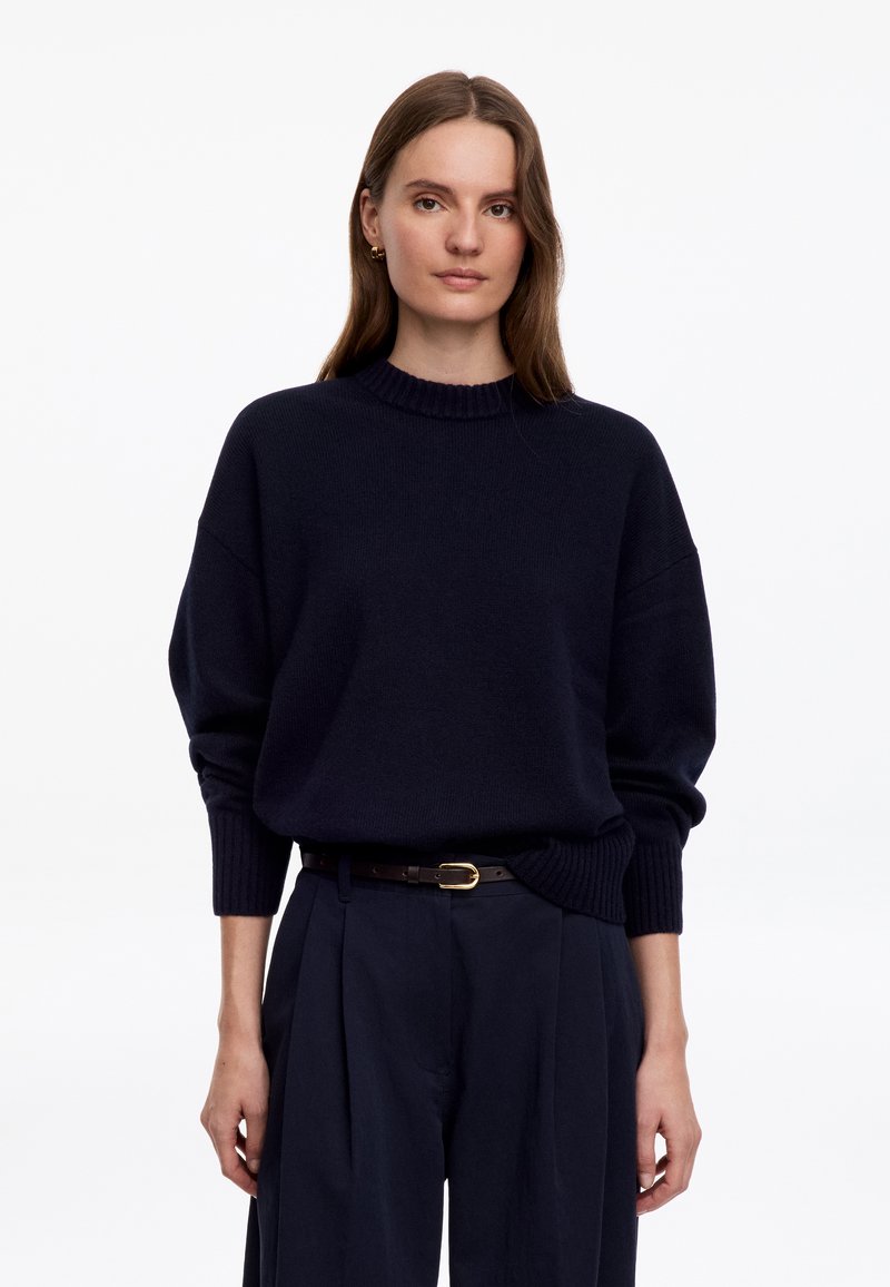 Femme aux cheveux bruns et lisses portant un pull bleu marine foncé et un pantalon assorti avec une ceinture noire, debout devant un fond blanc uni.