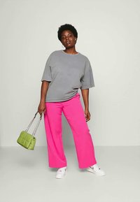 Kvinde iført en løs grå t-shirt, lys pink vidde ben bukser, hvide sneakers og holder en lille grøn håndtaske med kædestrop.