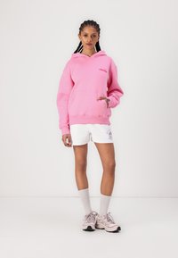 Rosa huvtröja med en framficka och logotyp, ihop med vita shorts med grafiska detaljer. Bärs med ljusa sneakers och ankelsockor.
