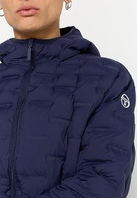 Granatowa kurtka puchowa z pikowanym wzorem i zapięciem na zamek. Posiada kaptur oraz haftowane logo na lewym ramieniu. Gładka faktura.