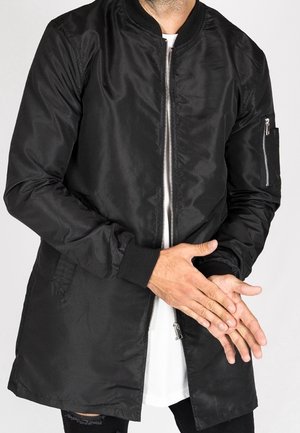 Homme portant une longue veste bomber noire avec une fermeture éclair, debout les mains jointes devant, sur une chemise blanche.