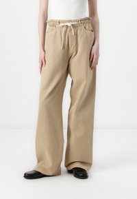 Beige vide benbukser med snor i livet, frontlommer og en avslappet passform. Glatt stoff med en subtil tekstur. Svarte sko synlige.