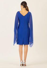 Königsblaues Kleid mit transparenten, fließenden Ärmeln und einer körperbetonten Silhouette. Verfügt über einen V-Ausschnitt am Rücken und einen verdeckten Reißverschluss. Silberne Riemchen-Heels.