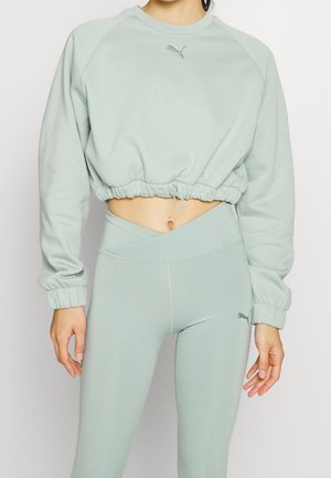 Frau trägt hellgrünes, kurz geschnittenes Sweatshirt und hoch taillierte Leggings mit Puma-Logos, zeigt den Bauch und steht mit entspannten Armen.
