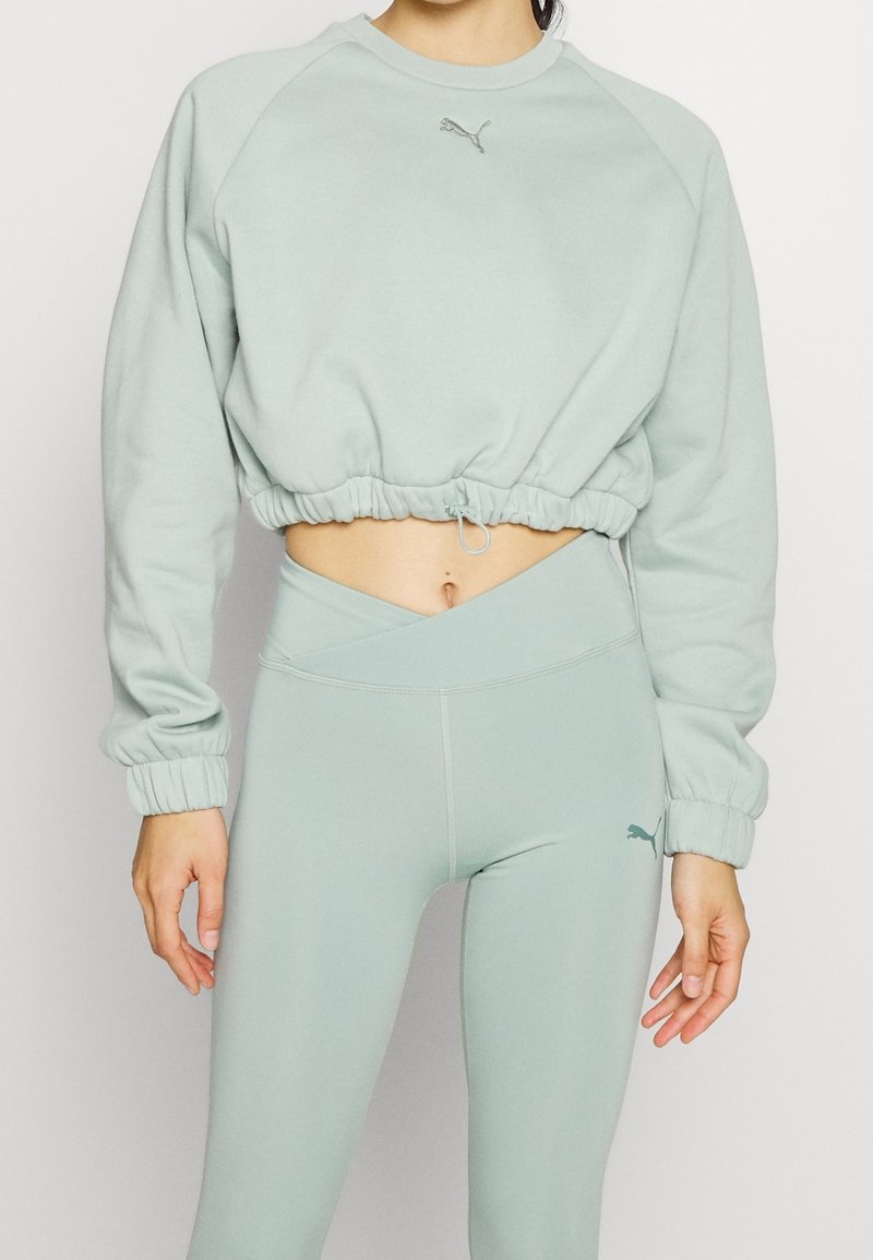 Femme portant un sweat-shirt court vert clair et des leggings taille haute avec des logos Puma, montrant le ventre et debout avec les bras détendus.
