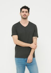 Άντρας με σκούρα μαλλιά και γένια, φορώντας γκρι σκούρο T-shirt με v-λαιμόκοψη και ανοιχτόχρωμο μπλε τζιν, στέκεται με το ένα χέρι να είναι σταυρωμένο πάνω από το άλλο.