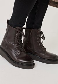 Barna bőr bokacsizma fűzővel és oldalsó cipzárral, textúrált díszítéssel és vastag fekete talppal, világos háttér előtt.
