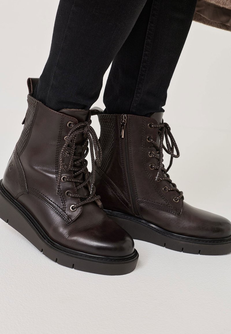 Barna bőr bokacsizma fűzővel és oldalsó cipzárral, textúrált díszítéssel és vastag fekete talppal, világos háttér előtt.