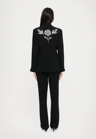 Costume noir sur mesure comprenant un blazer ajusté avec une broderie de rose et de feuille en argent au dos, associé à un pantalon droit.