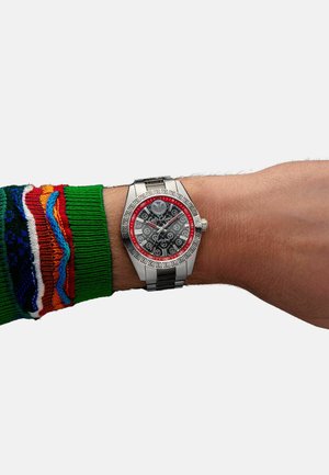 Silver watch with a black dial and red accents, featuring silver links and a patterned bezel. Worn on a wrist with a green, multicolored sweater.