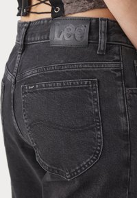 Black denim jeans featuring a textured finish, visible stitching, and a leather patch with "Lee" logo on the waistband and back pocket.