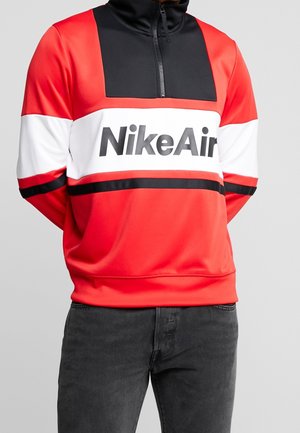 Man draagt een rode, witte en zwarte Nike Air trui met kwartritssluiting, staat met handen achter de rug en draagt donkere jeans.